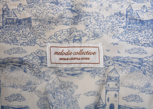 Close-up of a blue and white toile patterned fabric with a sewn-on brand label reading 'melodie collective SWIM & LIFESTYLE GOODS'.
