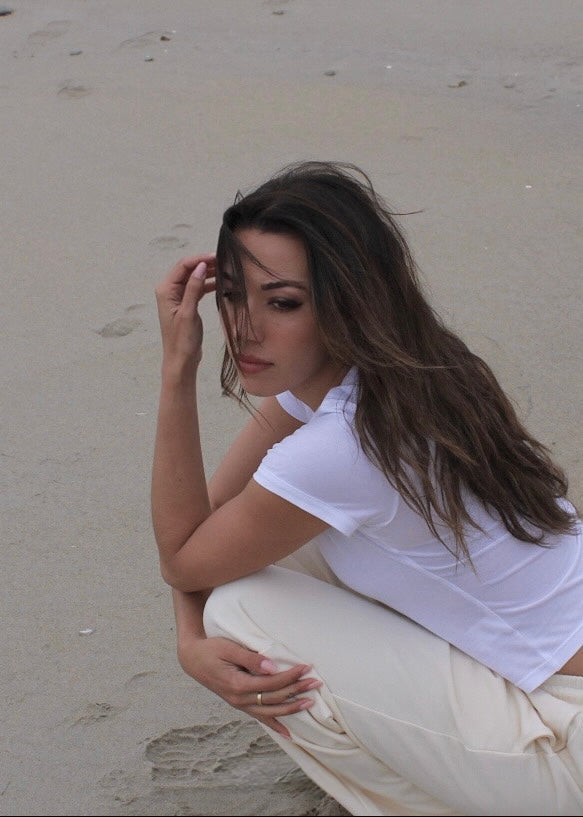 A woman with long dark hair crouches on a sandy beach, wearing a white short-sleeve shirt and cream-colored pants.