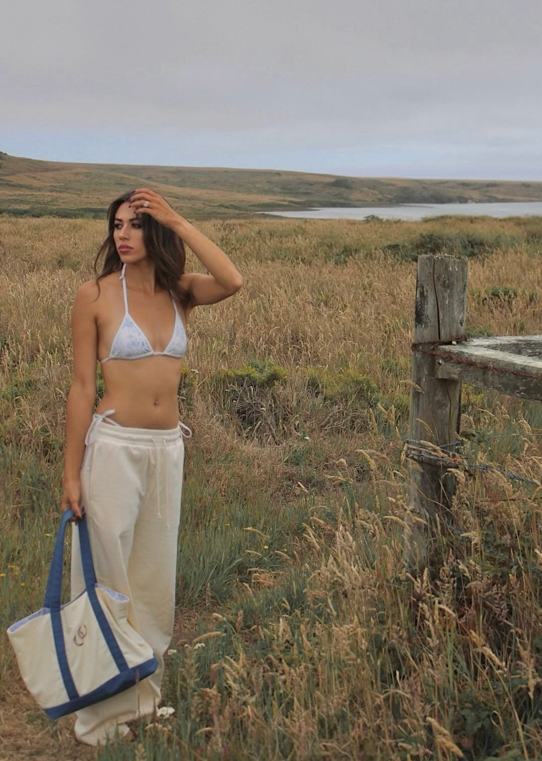 A woman stands in an open grassy landscape holding a cream canvas tote bag with blue handles. She is wearing a cream bikini top and loose cream pants, with a wooden fence and a body of water in the background under an overcast sky.