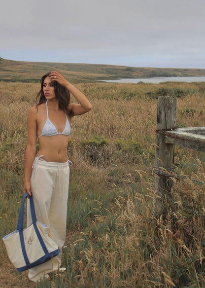 A woman stands in an open grassy landscape holding a cream canvas tote bag with blue handles. She is wearing a cream bikini top and loose cream pants, with a wooden fence and a body of water in the background under an overcast sky.