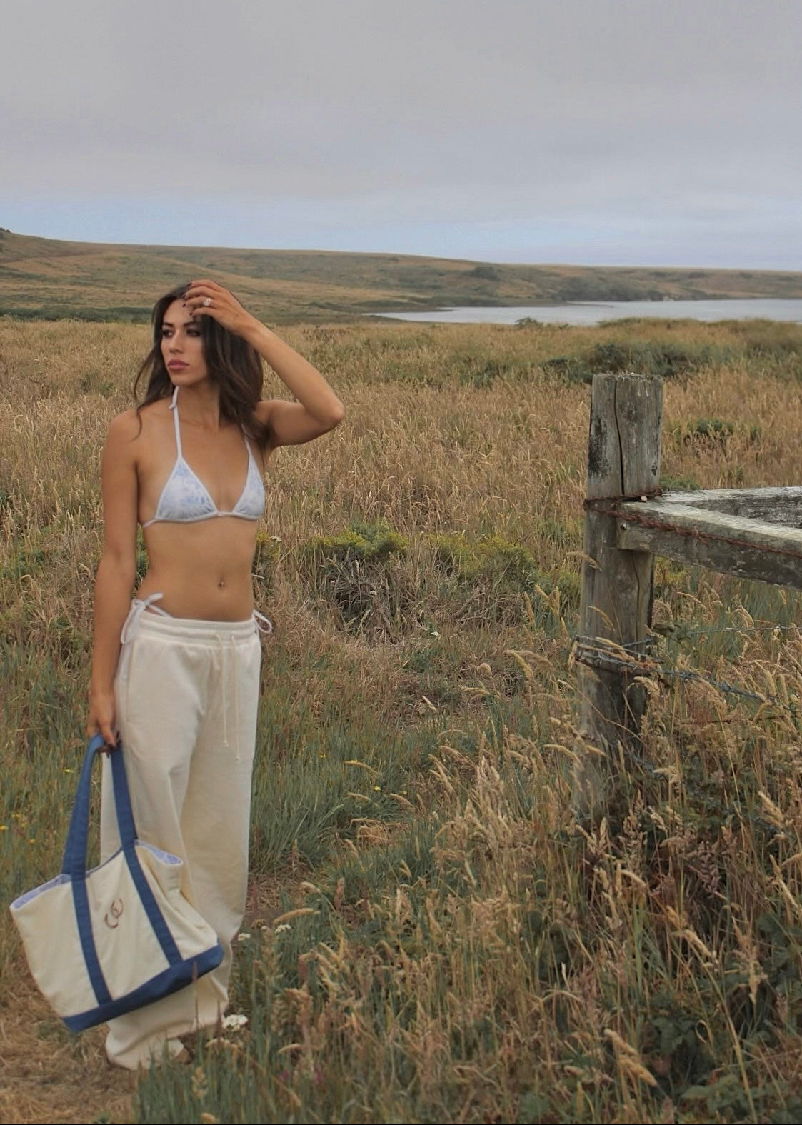 A woman stands in an open grassy landscape holding a cream canvas tote bag with blue handles. She is wearing a white bikini top and loose cream pants, with a wooden fence and a body of water in the background under an overcast sky.