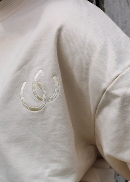 A close-up of a person wearing a cream-colored crewneck sweatshirt with a small embroidered logo on the chest, set against a weathered, peeling gray wall.