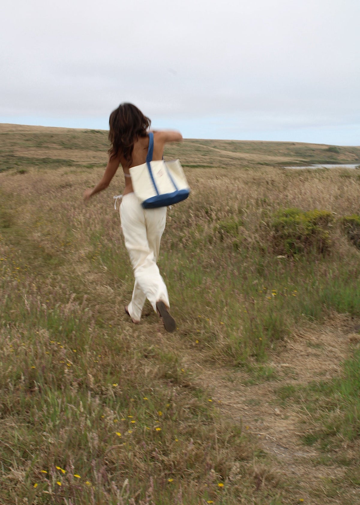 A woman walking outdoors on a grassy path carrying a large cream tote bag with blue handles and trim.