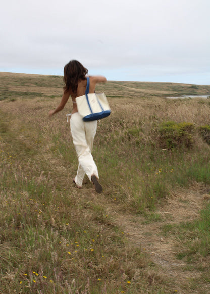 A woman walking outdoors on a grassy path carrying a large cream tote bag with blue handles and trim.