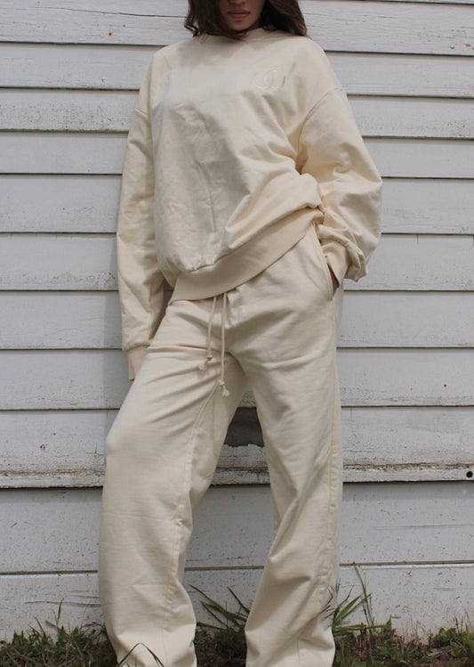 A woman wearing a matching cream-colored lounge set consisting of a long-sleeve sweatshirt and drawstring pants, standing against a white wooden siding wall.