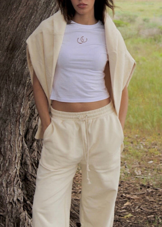 A woman stands outdoors next to a large tree trunk, wearing a white cropped tee with a small brown horseshoe logo, cream sweatpants, and a draped cream cardigan. The background shows a grassy field under a cloudy sky.