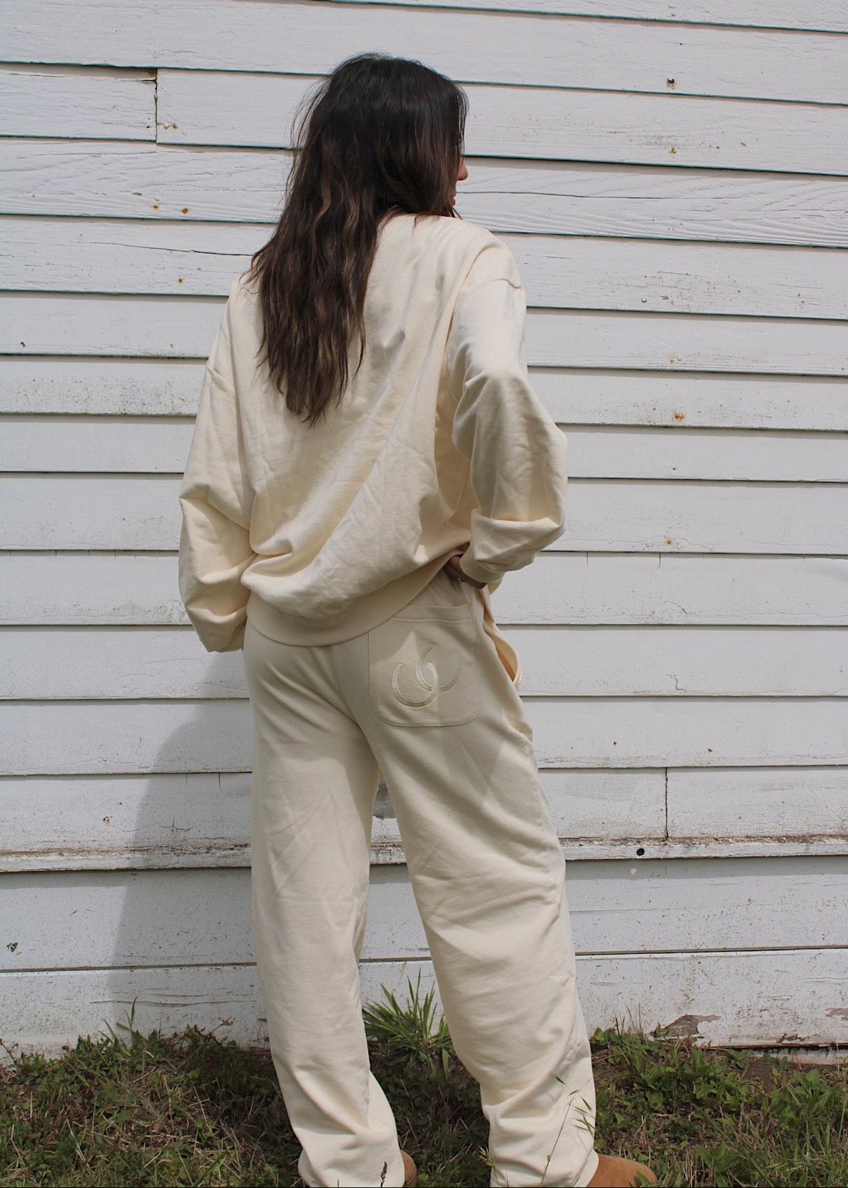 A person standing with their back to the camera, wearing a matching cream-colored lounge set consisting of a sweatshirt and sweatpants, set against a white wooden wall.
