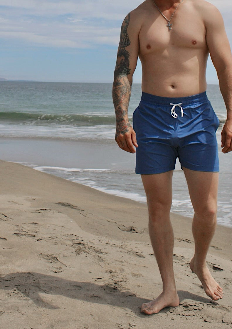 A man walking on a sandy beach wearing shiny blue swim trunks, a white cap, and sunglasses, with the ocean and cloudy sky in the background.