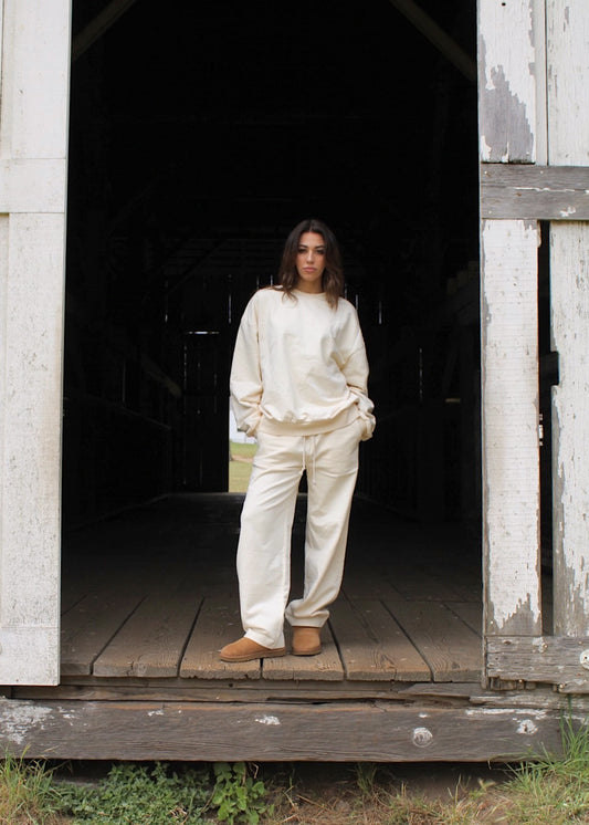 A woman stands in the doorway of a rustic white wooden barn, wearing a matching cream-colored lounge set consisting of a sweatshirt and wide-leg pants, paired with tan slip-on shoes.