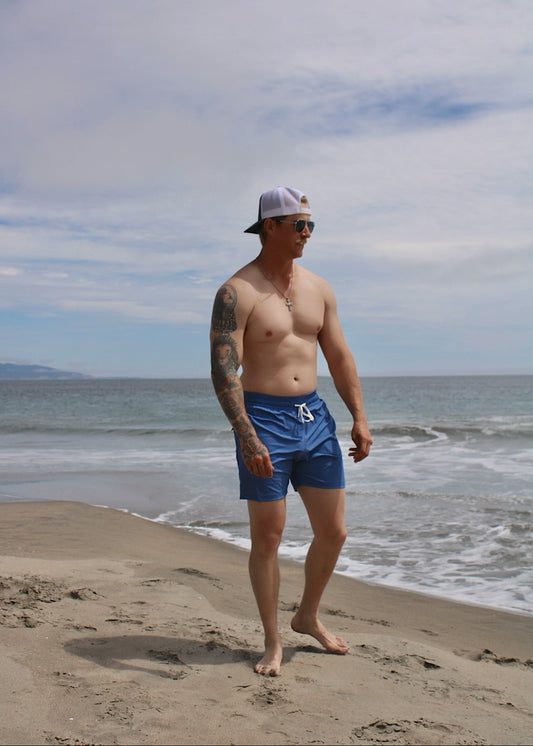 A shirtless man with tattoos stands on a sandy beach wearing blue swim trunks, a white cap, and sunglasses, with the ocean and cloudy sky behind him.