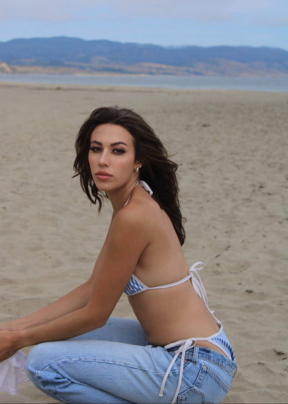 A woman crouching on a sandy beach looking at the camera and wearing a blue and cream striped bikini, jeans, and holding a shirt.