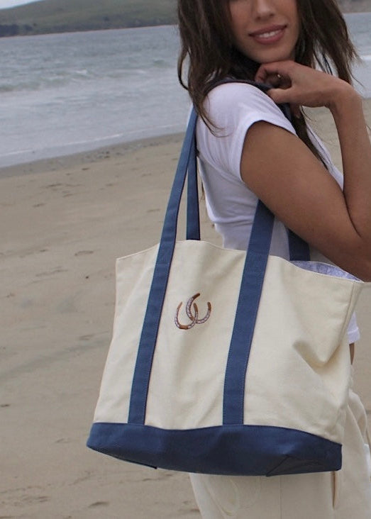 A woman on a beach carrying a large cream canvas tote bag with navy blue straps, bottom trim, and a small embroidered logo on the front.