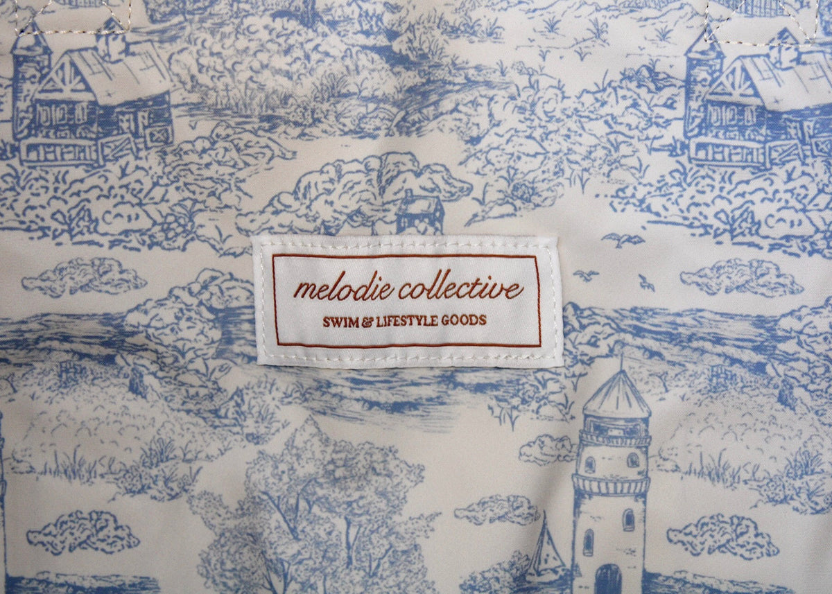 Close-up of a blue and white toile patterned fabric with a sewn-on brand label reading 'melodie collective SWIM & LIFESTYLE GOODS'.