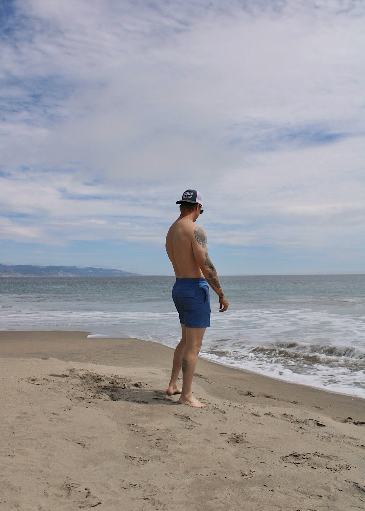 A man standing on the beach looking at the ocean wearing blue swim trunks and a blue cap.