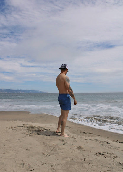 A man standing on the beach looking at the ocean wearing blue swim trunks and a blue cap.