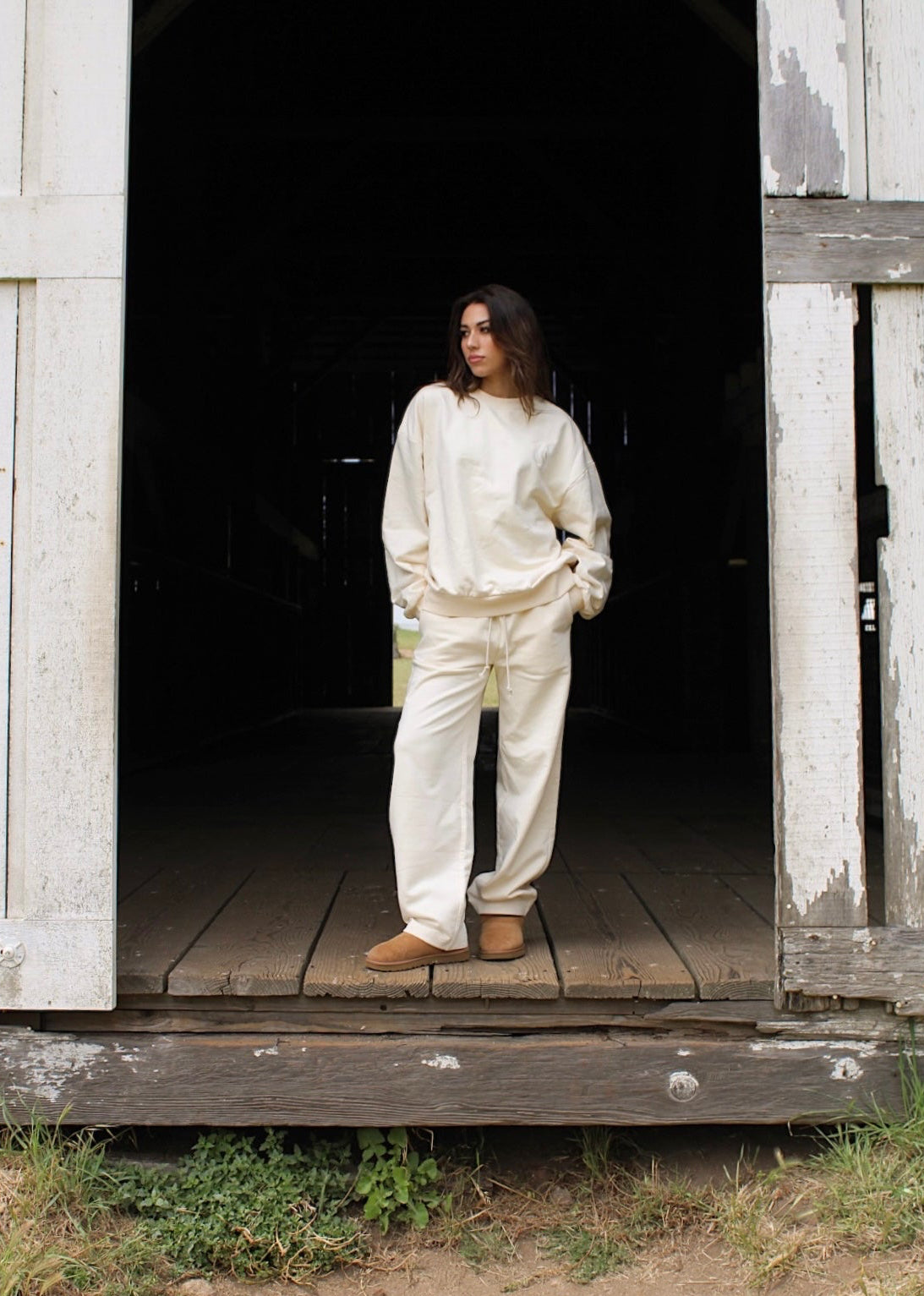 A model wearing a cream-colored lounge set consisting of a sweatshirt and matching sweatpants, standing in the doorway of a rustic wooden barn.