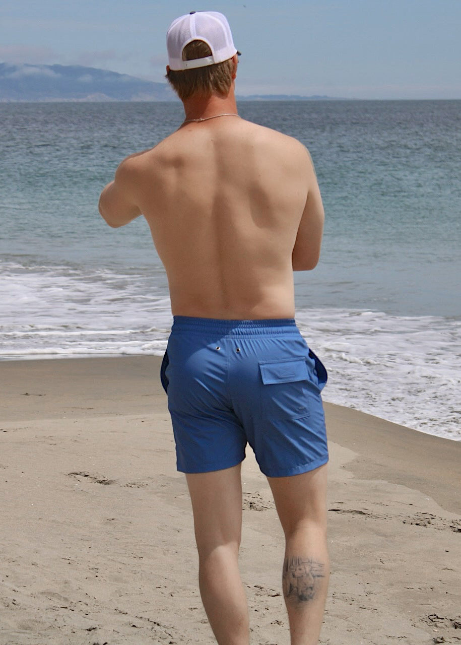A view from behind of a man standing on the beach wearing blue swim trunks and a white cap.