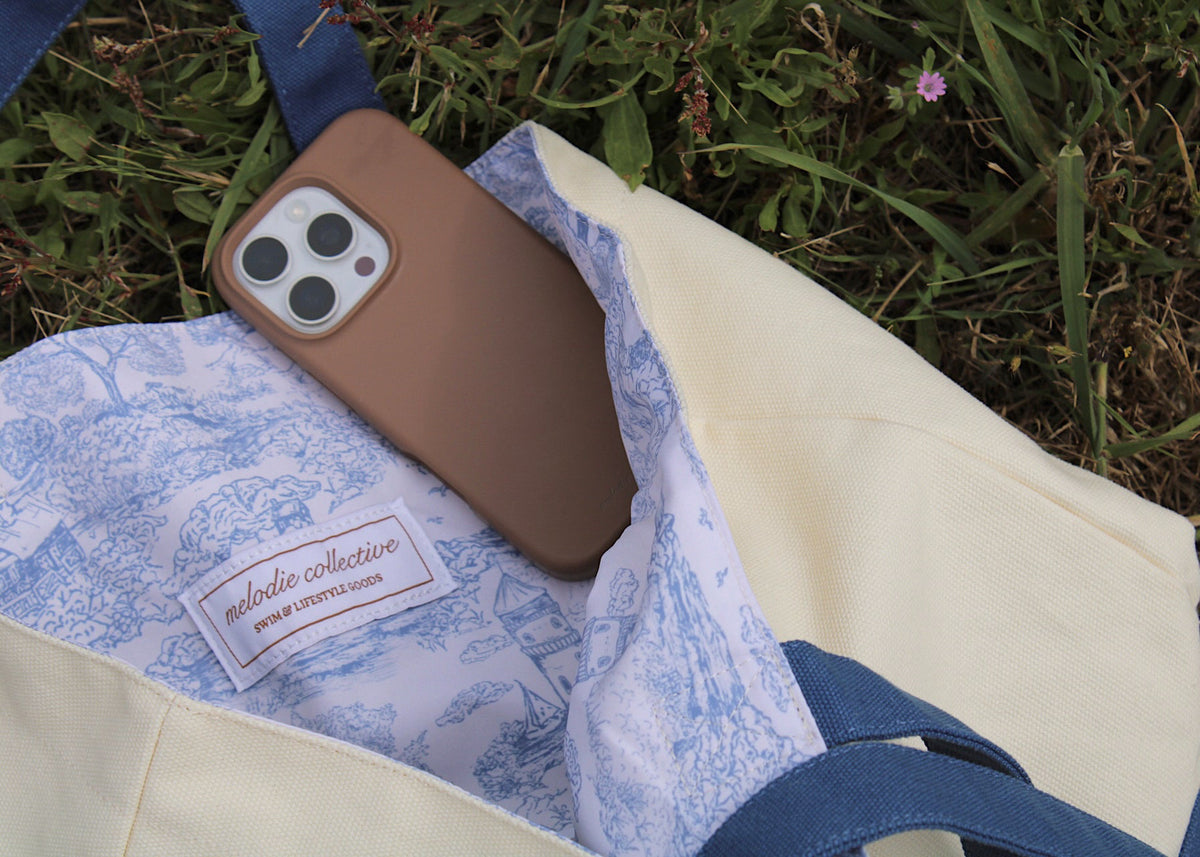 An open cream canvas tote bag with blue handles and a blue-and-white toile patterned lining, shown outdoors on grass with a brown smartphone resting inside. A sewn label reads Melodie Collective.