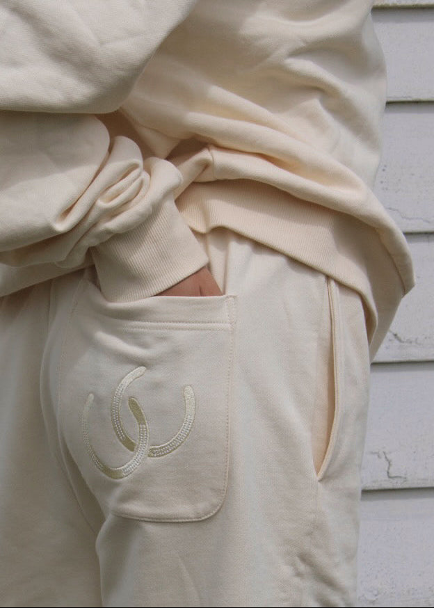Close-up of a person wearing cream-colored sweatpants with an embroidered interlocking horseshoe logo on the pocket, paired with a matching cream top, set against a white wooden wall.