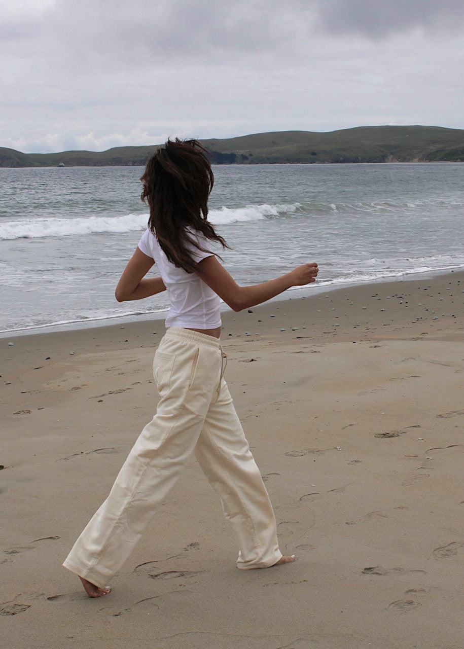 A woman walks barefoot on a sandy beach from behind, wearing a white cropped t-shirt and cream wide-leg trousers, with the ocean and green hills in the background under an overcast sky.