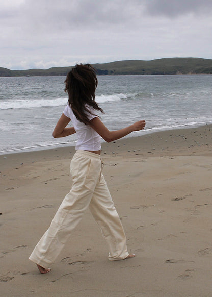 A woman walks barefoot on a sandy beach from behind, wearing a white cropped t-shirt and cream wide-leg trousers, with the ocean and green hills in the background under an overcast sky.