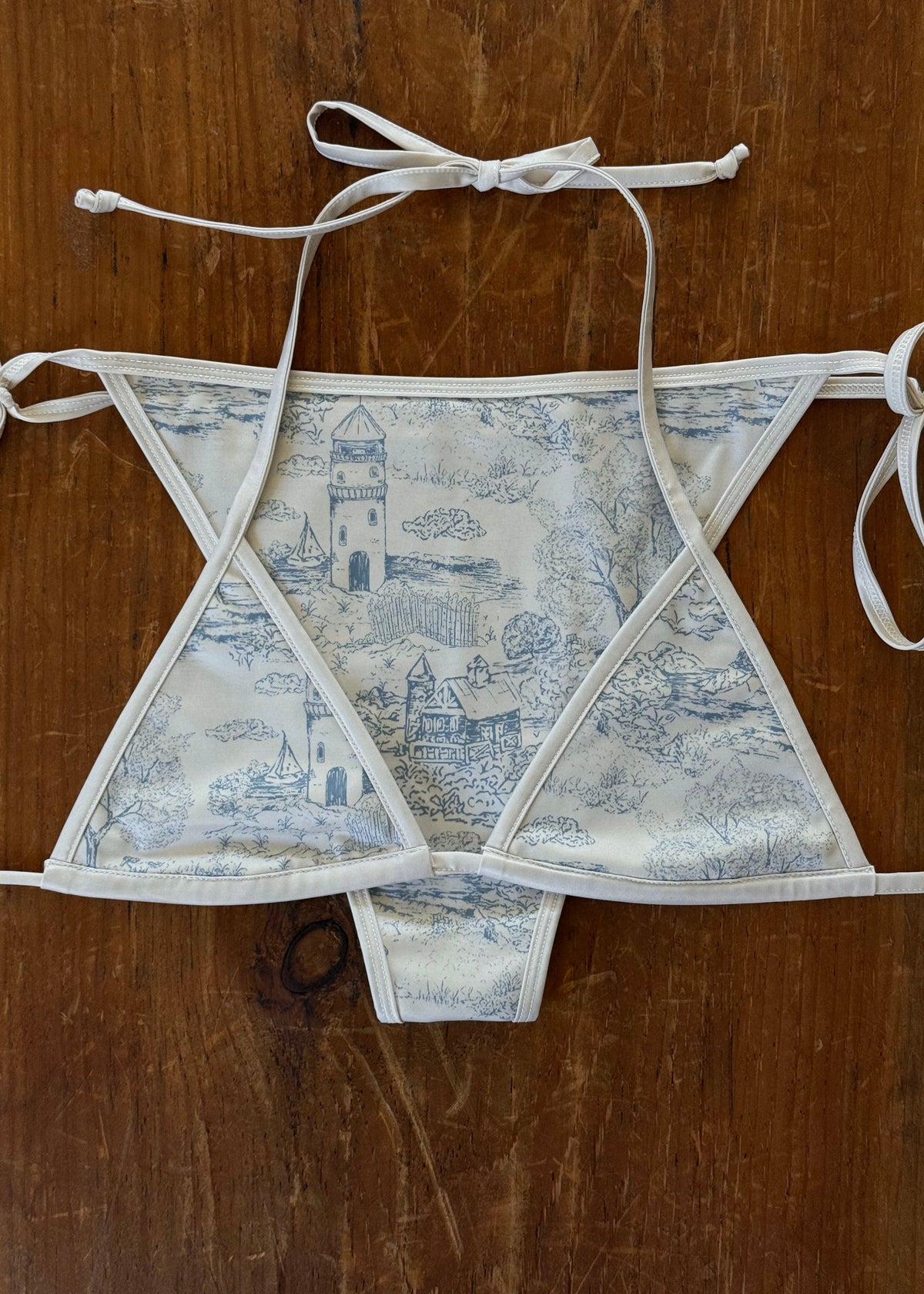 A structured triangle bikini top with a blue and white toile pattern, laid flat on a dark brown wooden surface.