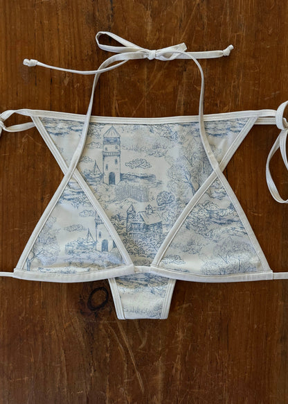 A structured triangle bikini top with a blue and white toile pattern, laid flat on a dark brown wooden surface.