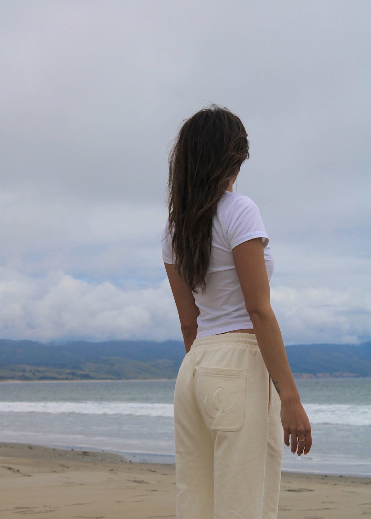 A woman with long dark hair stands on a sandy beach facing the ocean, wearing a white short-sleeve t-shirt and cream-colored wide-leg pants.