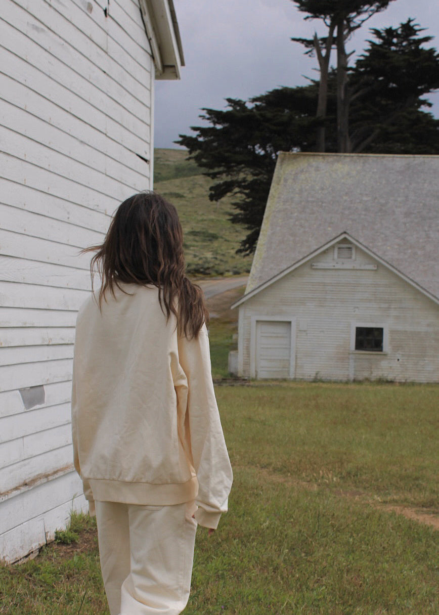 A person with long dark hair stands outdoors in front of a white wooden building, wearing a matching cream-colored sweatshirt and sweatpants set. Another white building and grassy field are visible in the background.