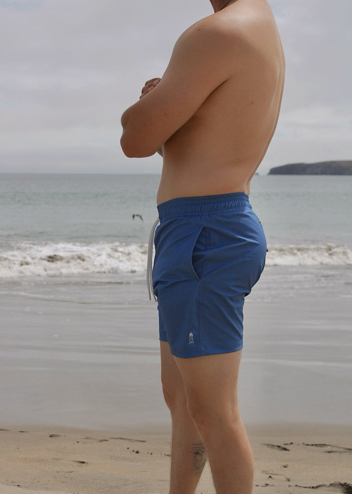 A side profile of a man standing on the beach wearing blue swim trunks with lighthouse embroidery.