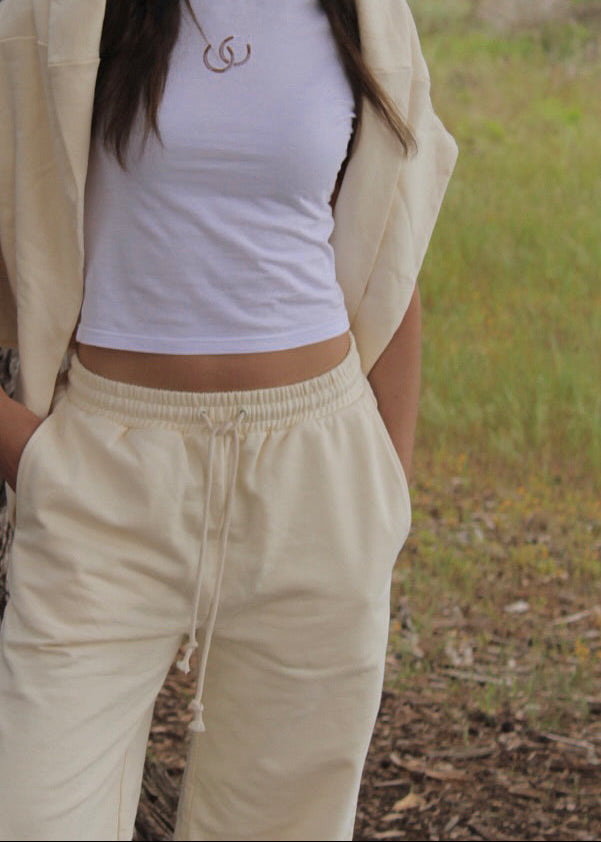 A person outdoors wearing a white cropped top, cream drawstring sweatpants, and a cream open-front jacket, with a small gold necklace visible.